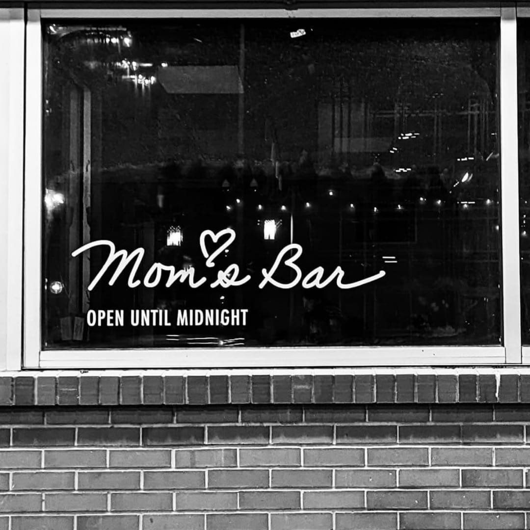 A window with the words 'Mom's Bar Open Until Midnight" on it in white. 