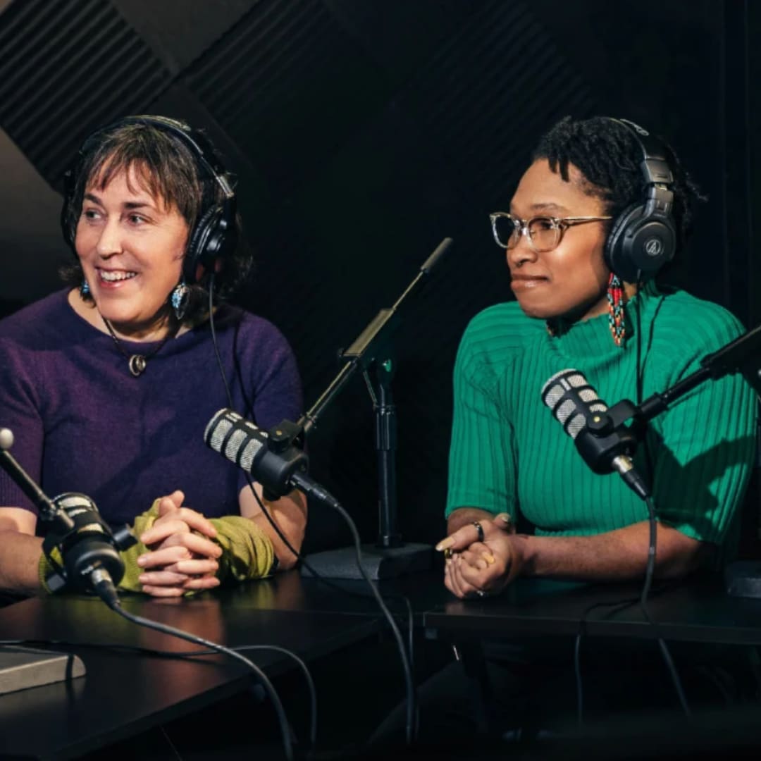 Two woman with headphones on, sitting near microphones. 