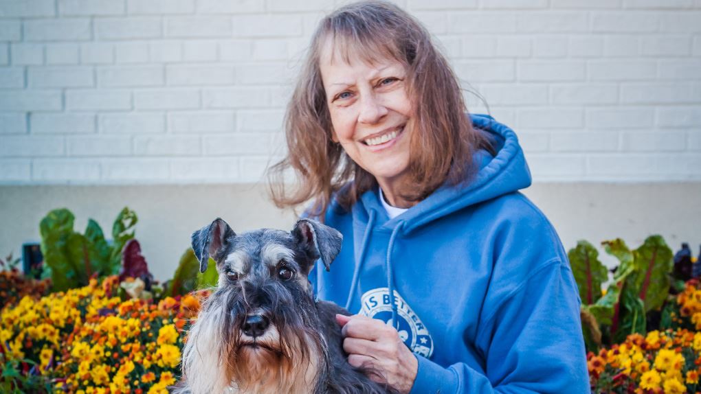 Dr. Linda Sullivan wearing a blue sweatshirt sitting in front of yellow flowers with her dog on her lap