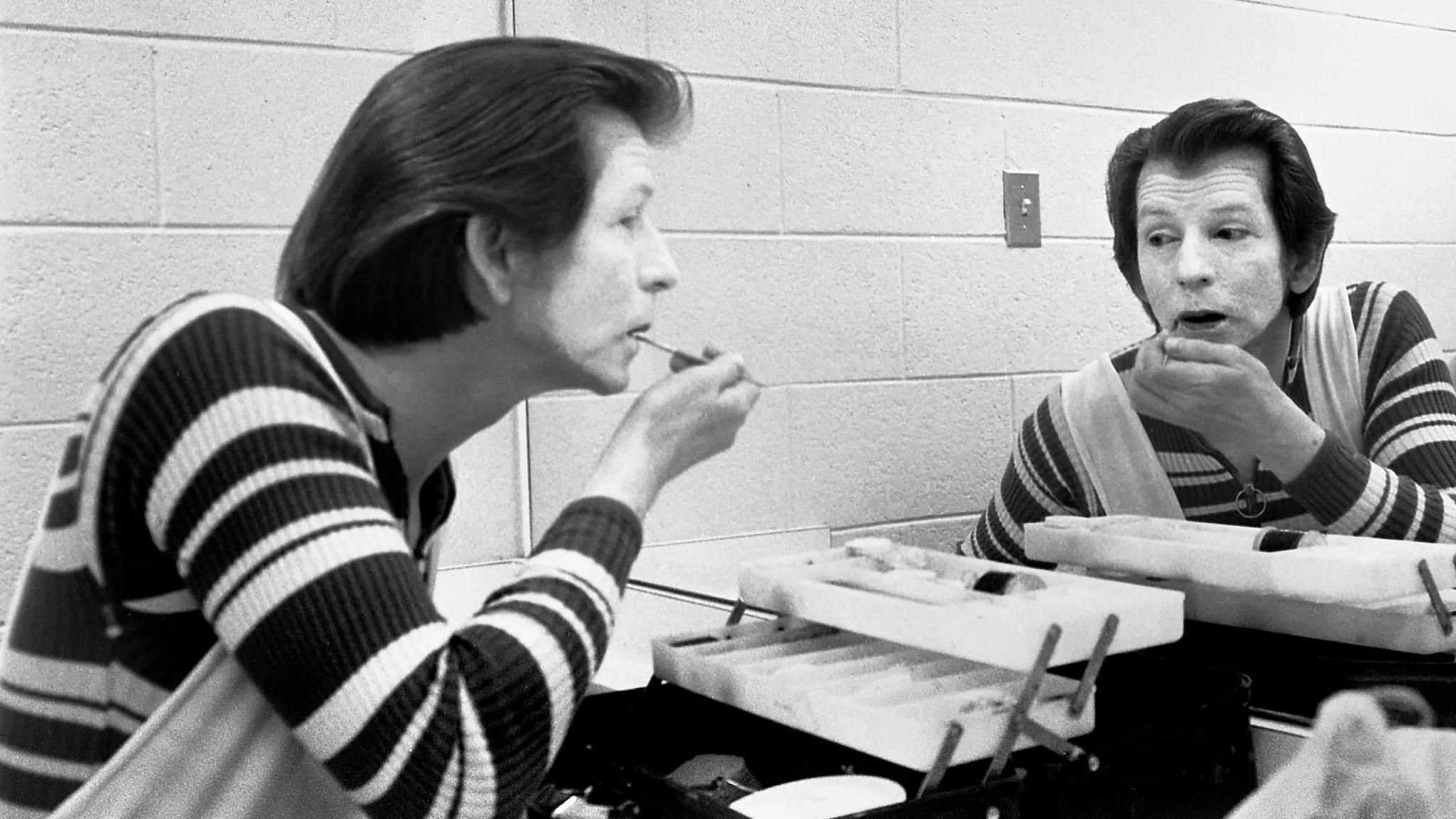 Black-and-white photo of mime master Reid Gilbert applying makeup while looking at himself in a dressing room mirror.