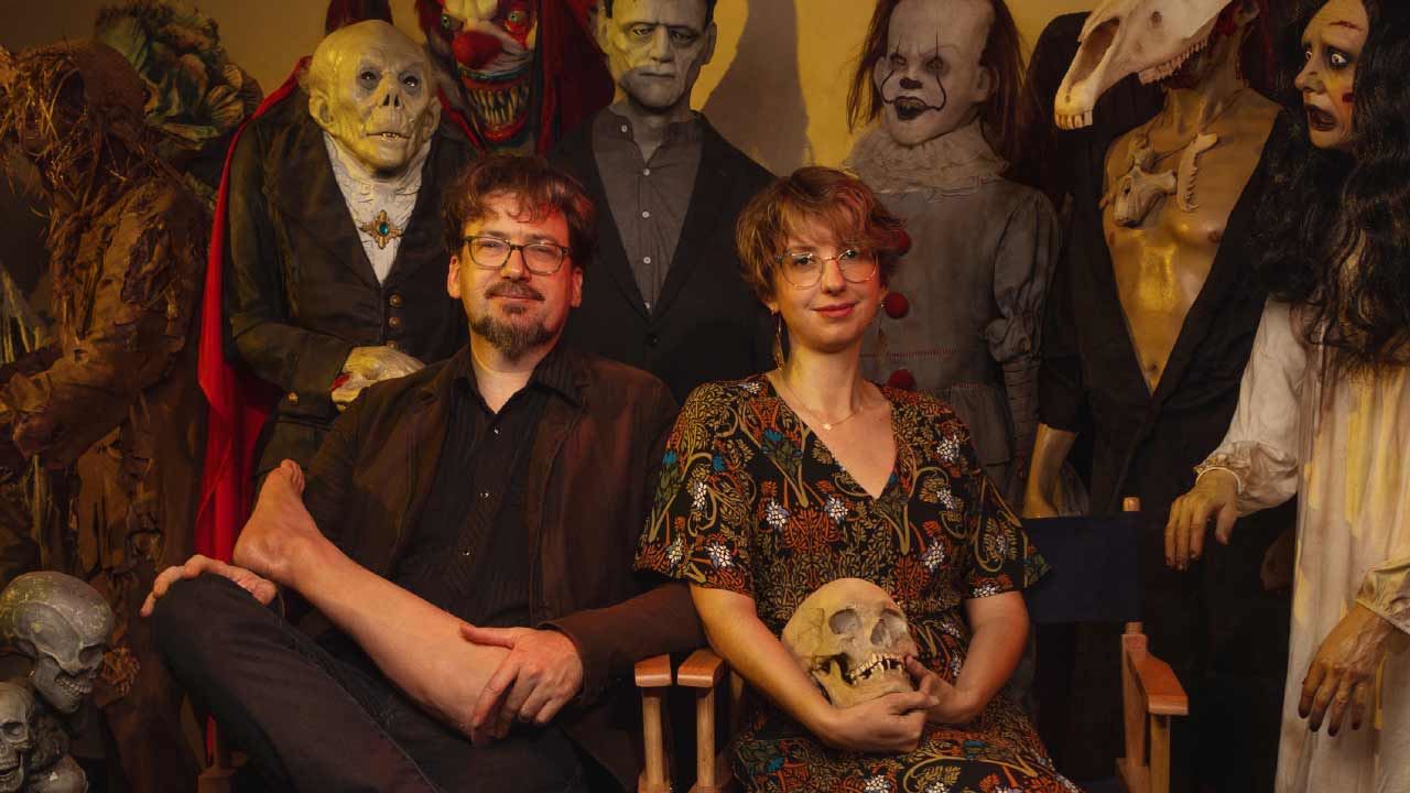 Dapper Cadaver owners are sitting side by side in front of some props holding an artificial leg and skull