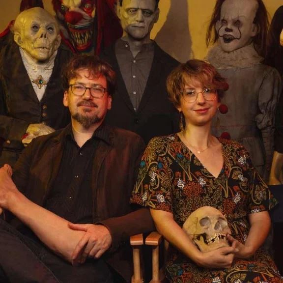 a man and a woman sitting in director chairs, he's holding a fake leg, she's holding a fake skull. There are three life-size mannequins (two zombies, one It clown) standing behind them