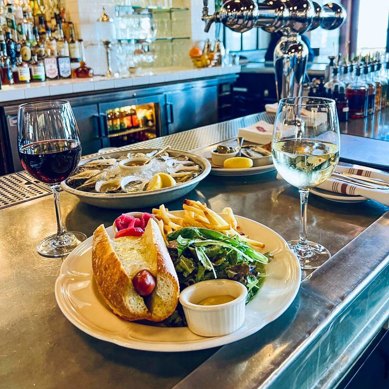 A plate with a hot dog, fries and greens sits next to two glasses of wine and a plate of oysters and lemon on a bar.