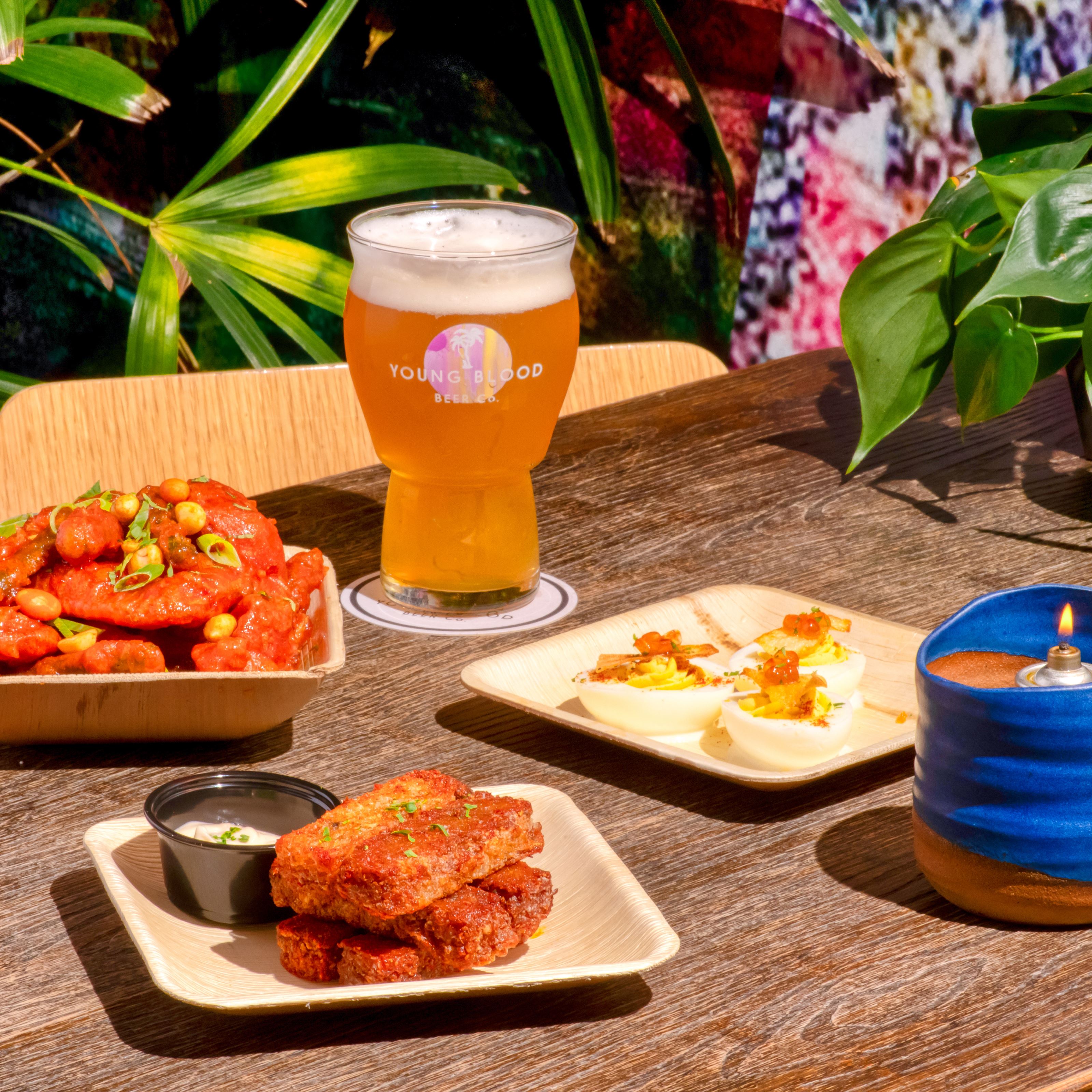 A wooden table is laid out with a beer, plates of snacks, and a blue candle. The background is flanked with green plants.