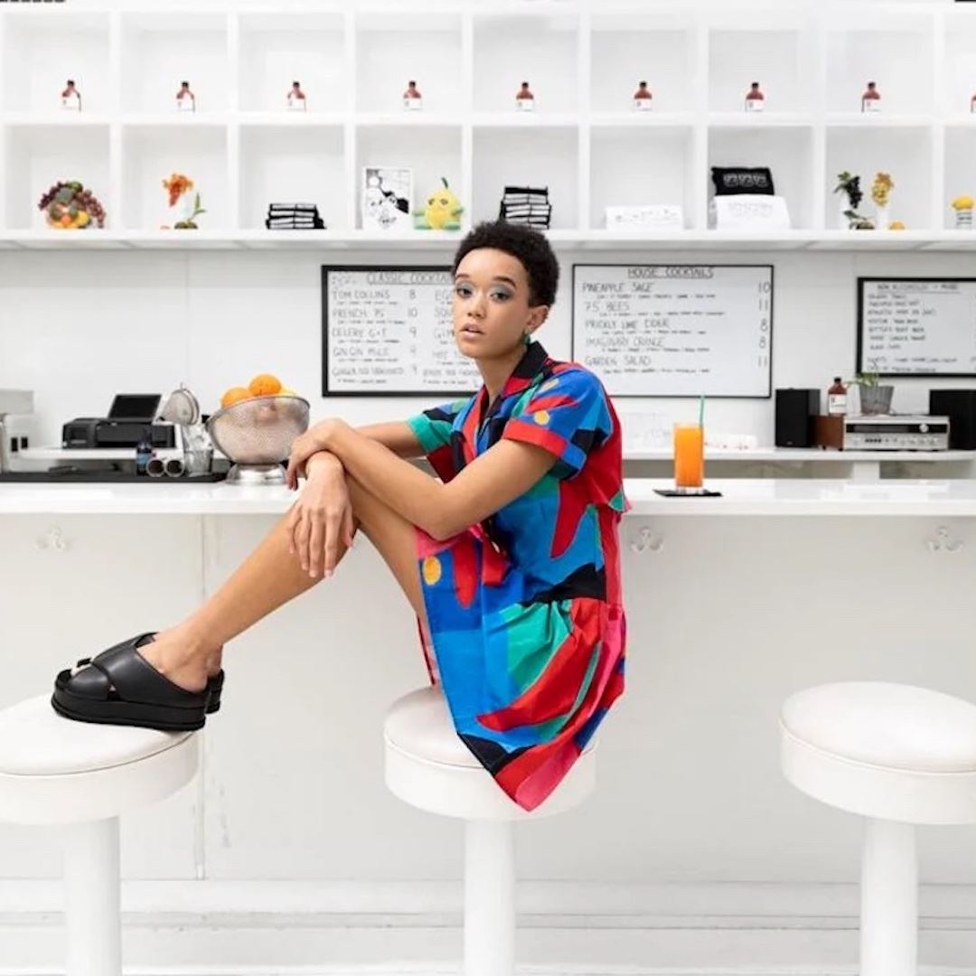 Woman sitting with her feet on the stool next to her in a brightly colored dress and black sandals