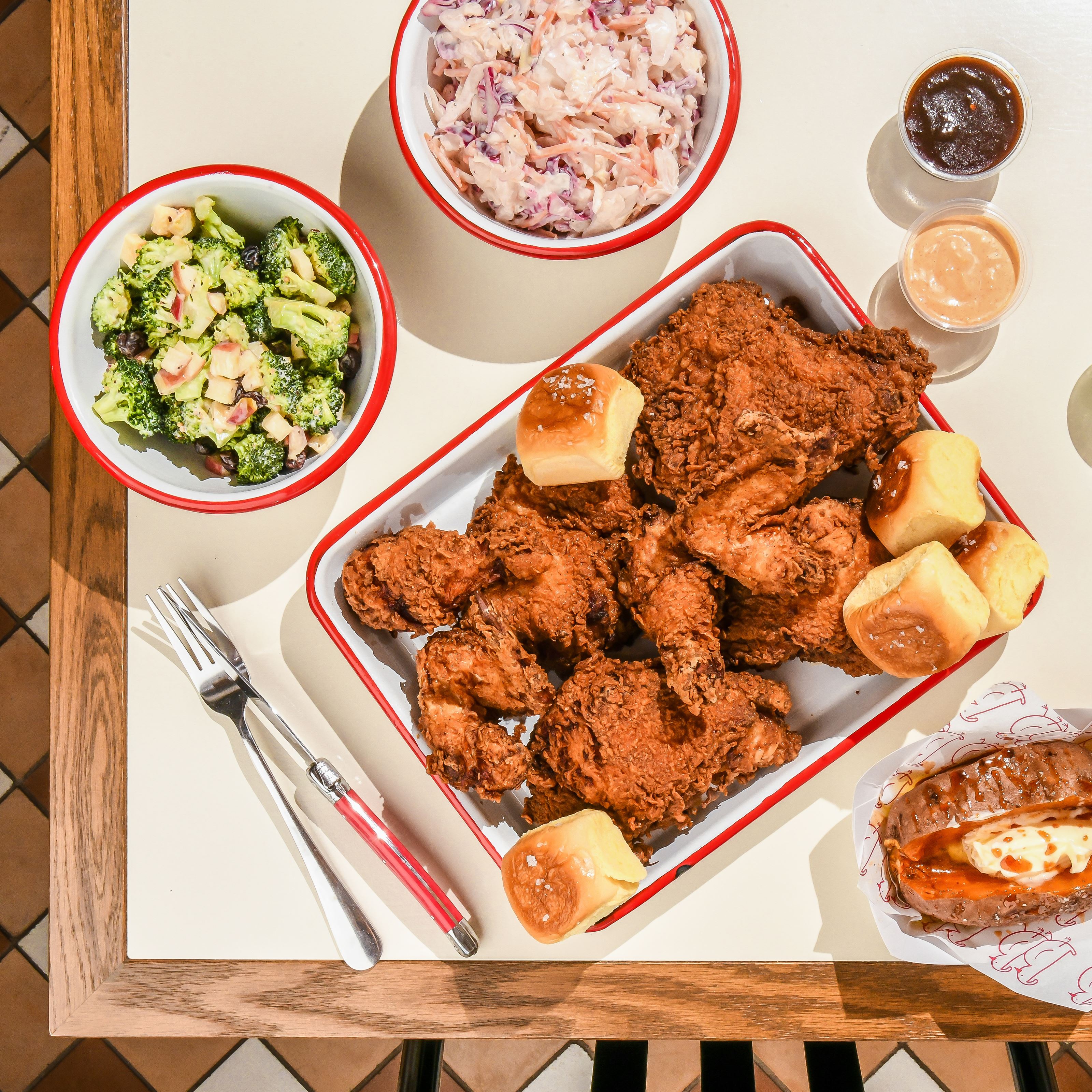 A white table displays a chicken dinner at Butterbird, including a platter of fried chicken and dinner rolls, a bowl of broccoli, a bowl of slaw, a hot honey sweet potato and containers of dipping sauce.