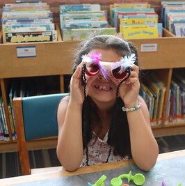 Young girl poses for picture in front of stacks of library books while sitting at a table and looking through handmade paper binoculars.