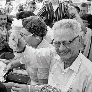 Black and white photo of man holding up an ice cream cone while winking, amidst a crowd of people.