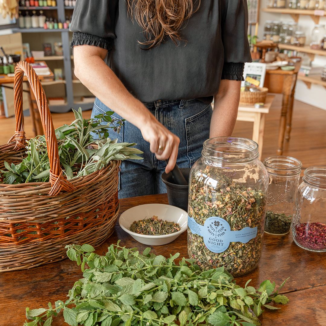 A woman wearing a gray shirt and a pair of blue denim jeans sprinkles dried herbs into a white bowl. Around her on a wooden table are a basket of fresh herbs, a pile of fresh herbs, a large jar of herbal tea and two smaller jars of herbs.