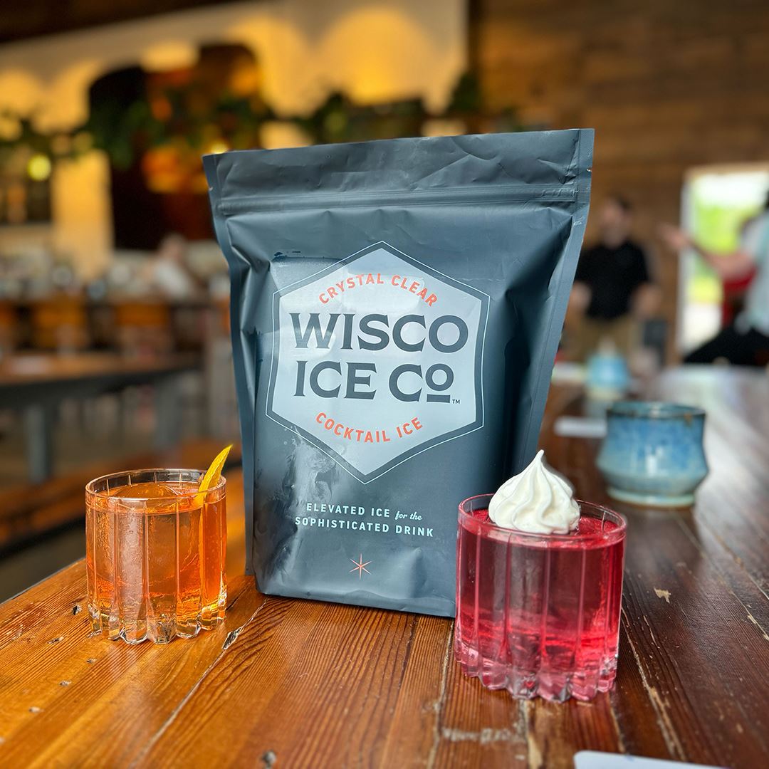 A navy bag of Wisco Ice Co. ice sits in between an orange cocktail and a red cocktail with a whipped cream dollop.