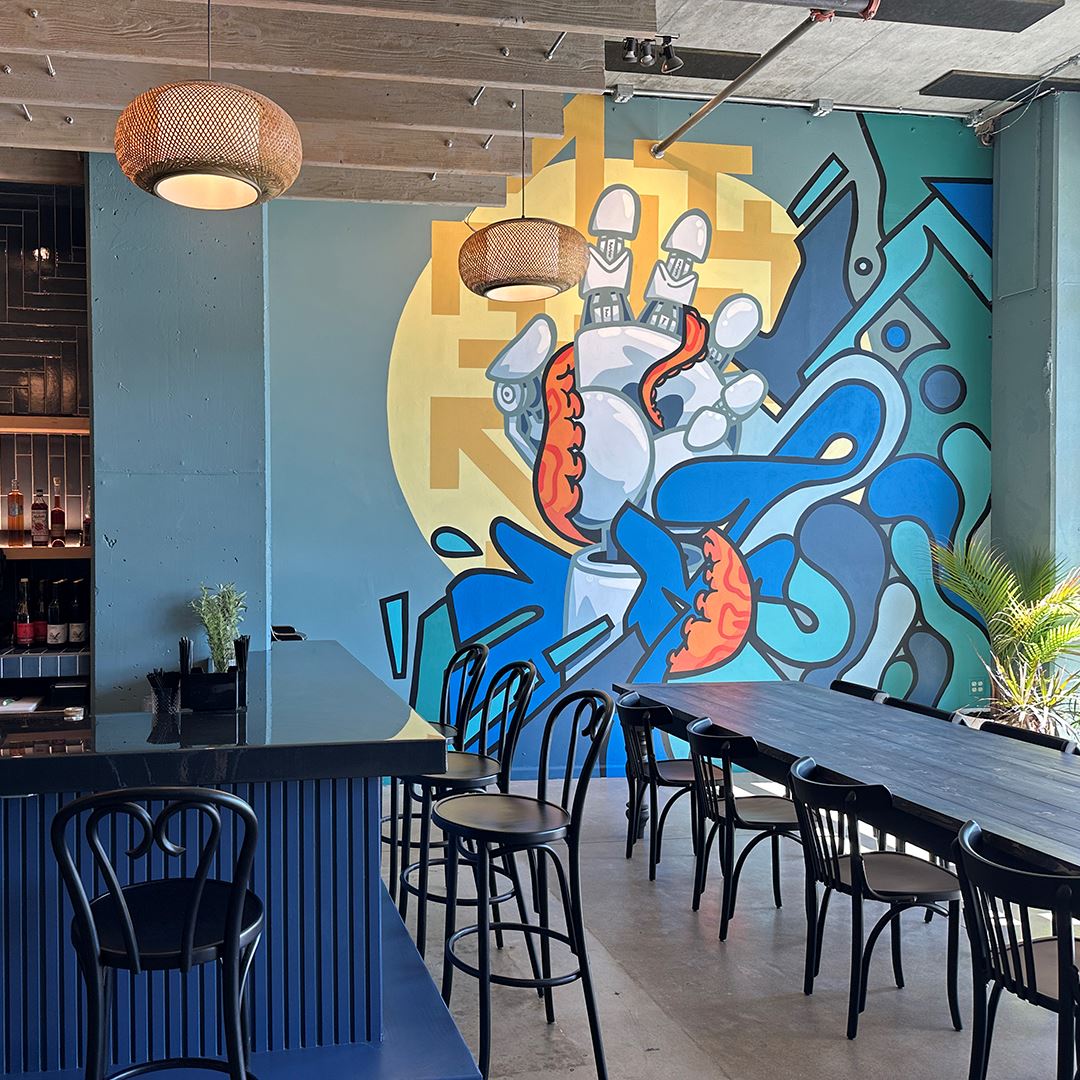 A blue, orange and yellow mural is the main focus of this photo, showing a robot hand reaching out of the waves with an orange octopus tentacle wrapped around it. In the foreground of the mural, a long black table and chairs stretches into view, next to a navy cocktail bar.