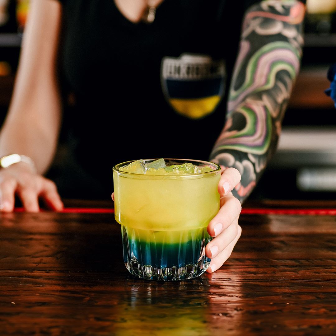 A blue and yellow cocktail is in the forefront of this image, resting on a dark wooden bar. An arm with a colorful tattoo sleeve is setting the drink on the bar.