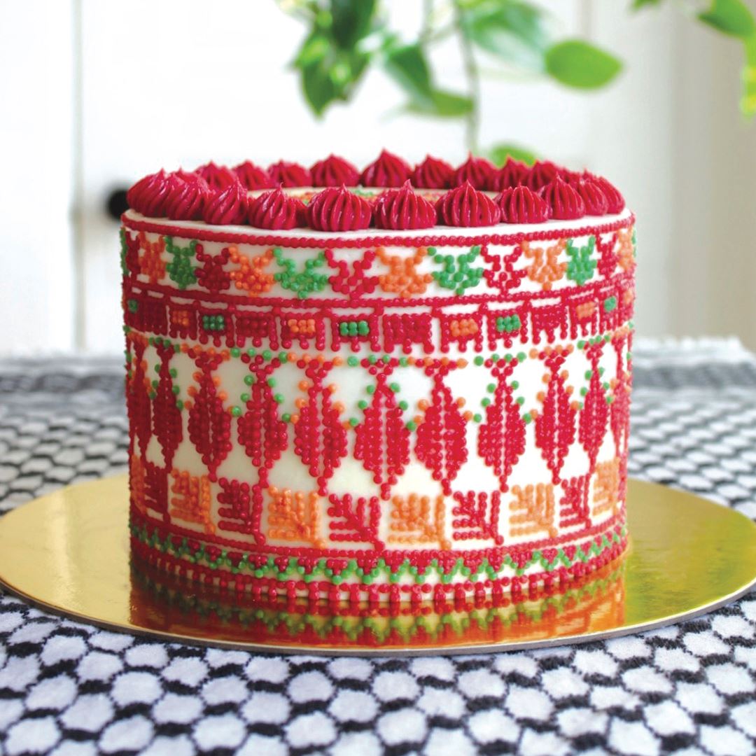 A cake is displayed on a golden platter. The white cake is decorated with intricate dot designs in red, green and orange.