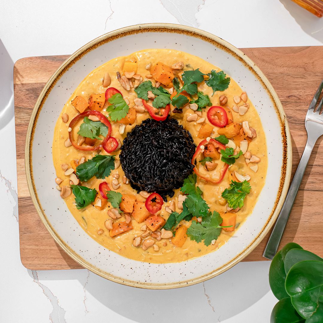 A wooden cutting board is set on a white marble counter. On the wooden cutting board sits a vibrant orange, red and green dish with black rice in the center.