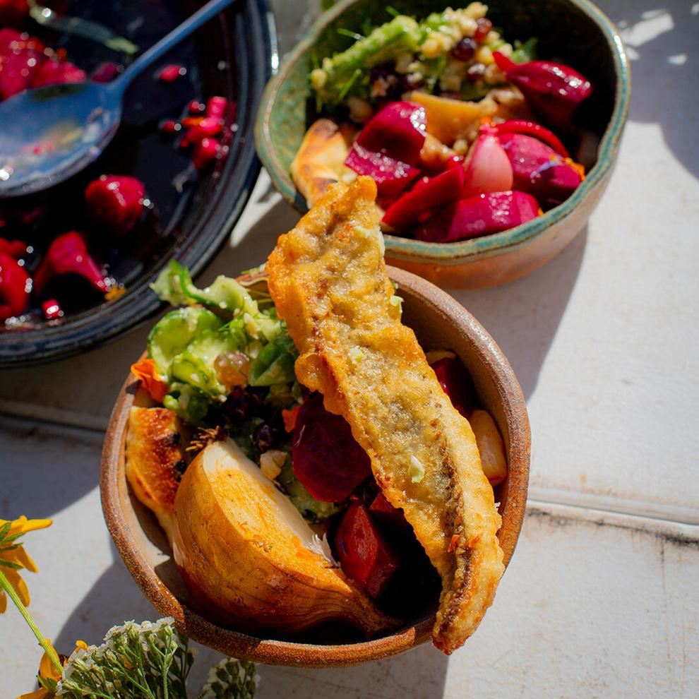 On a sunlit white surface, a brown pottery bowl displays a fried fish dish. In the background, another pottery bowl holds bright red produce and grain salad.