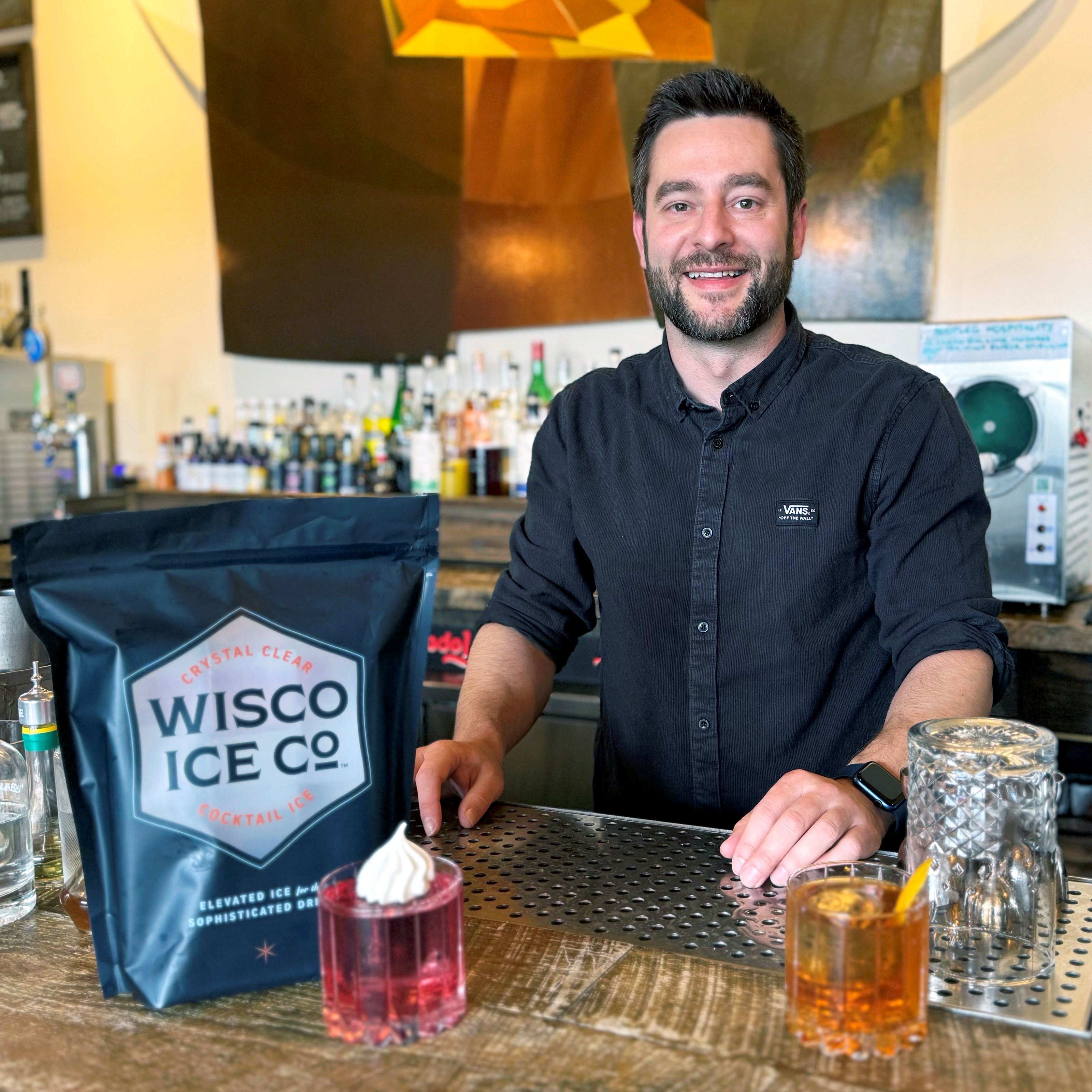 A man in a dark grey button down shirt stands behind a bar, where a bag labeled "Wisco Ice Co." and two drinks sit atop.