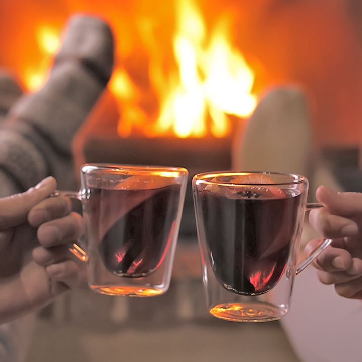 Two hands holding glasses on amber liquid toast each other in front of a fireplace.