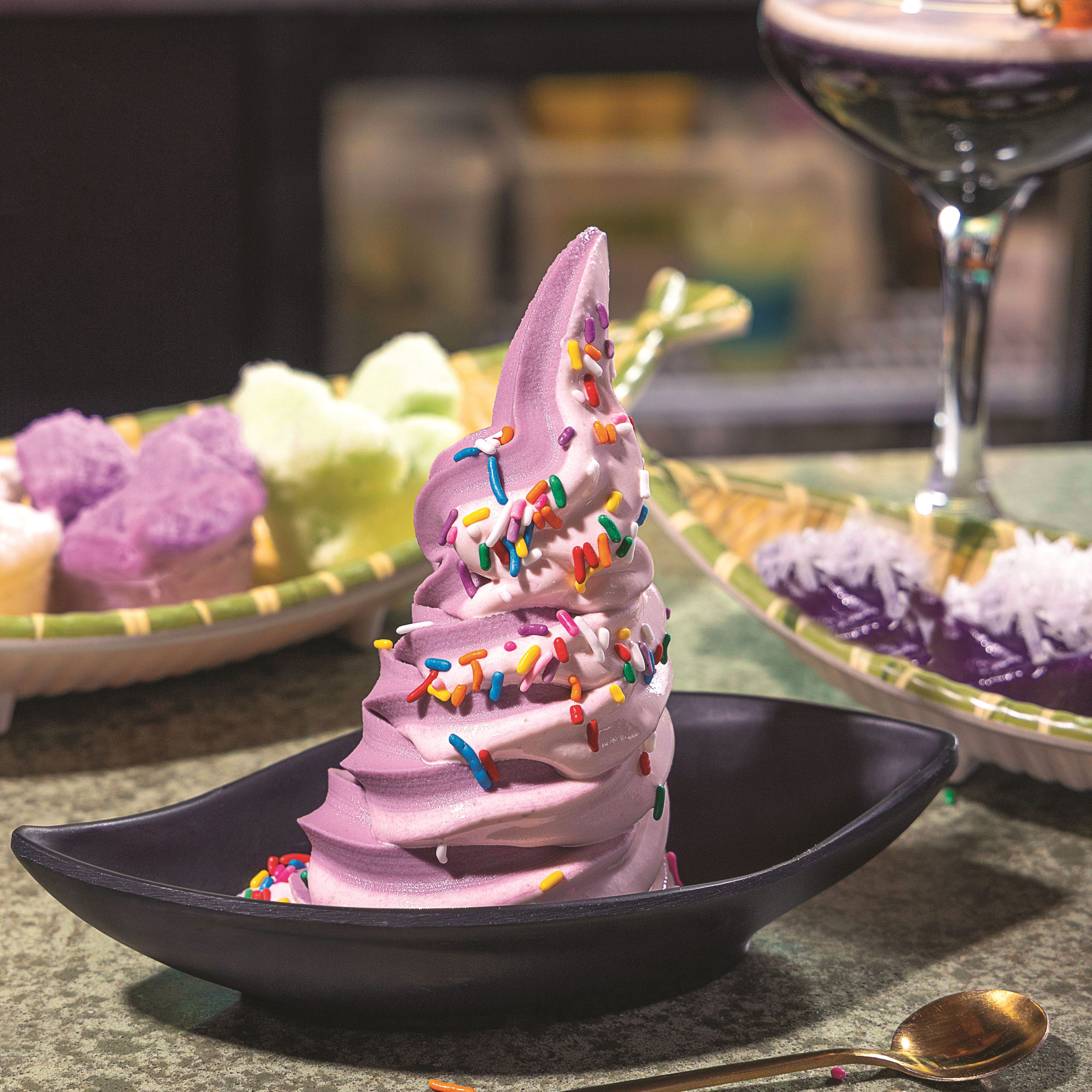 A swirl of purple soft serve ice cream emerges from a black dish. In the background, fish-shaped baskets display Thai rice cakes and a martini glass holds a purple-colored cocktail.