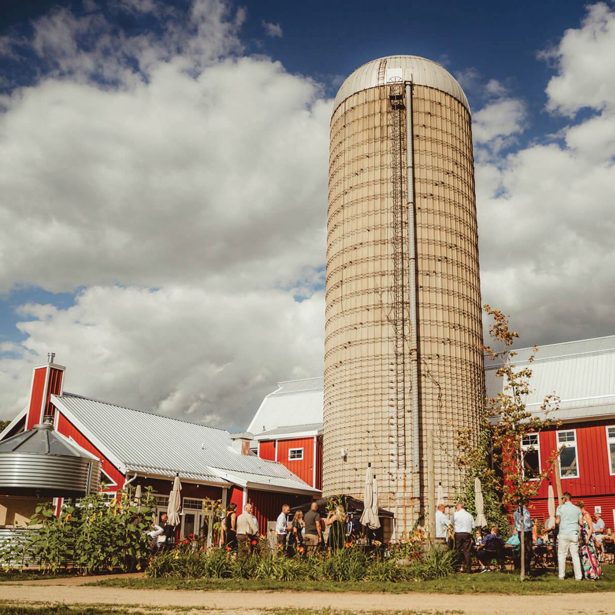 A brown farm silo rises from a bright red barn into a blue sky with puffy white clouds. People linger around the barn.