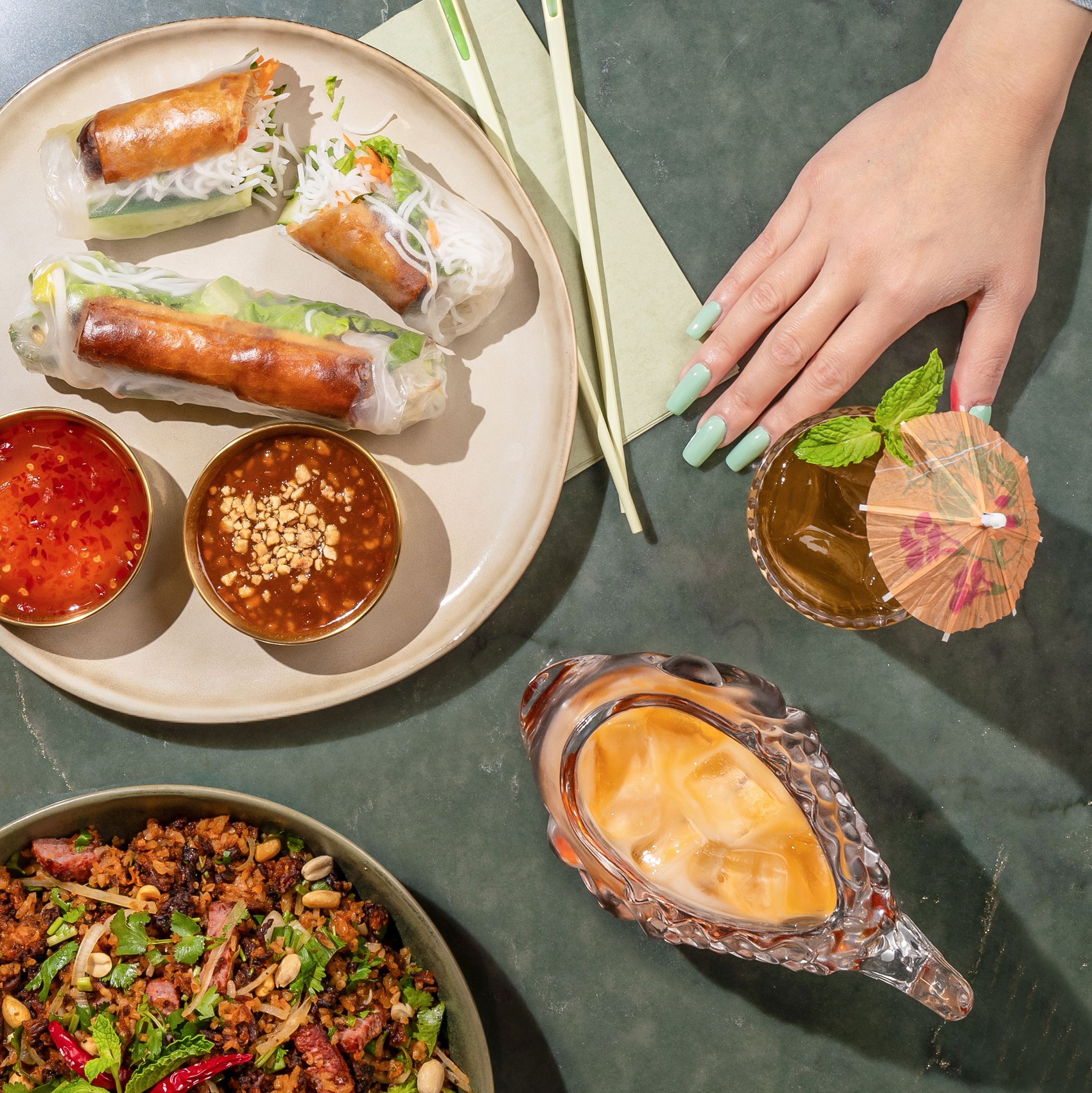 On gray/green granite countertop, a beige plate displays spring rolls and two dipping sauces, a bowl displays a fried rice dish, a glass vessel holds an iced coffee drink and a glass holds a cocktail with a cocktail umbrella. A hand with fingernails painted light blue reaches into the photo.