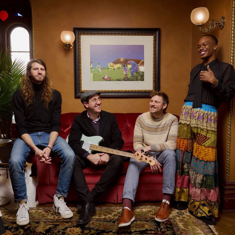 Members of Spectaculous pose on a couch with a piano and bass guitar. 
