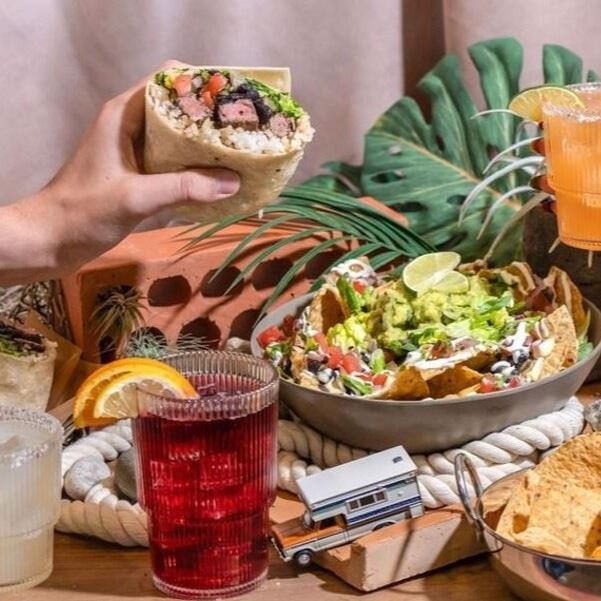 A hand holding a burrito above a table full of food.
