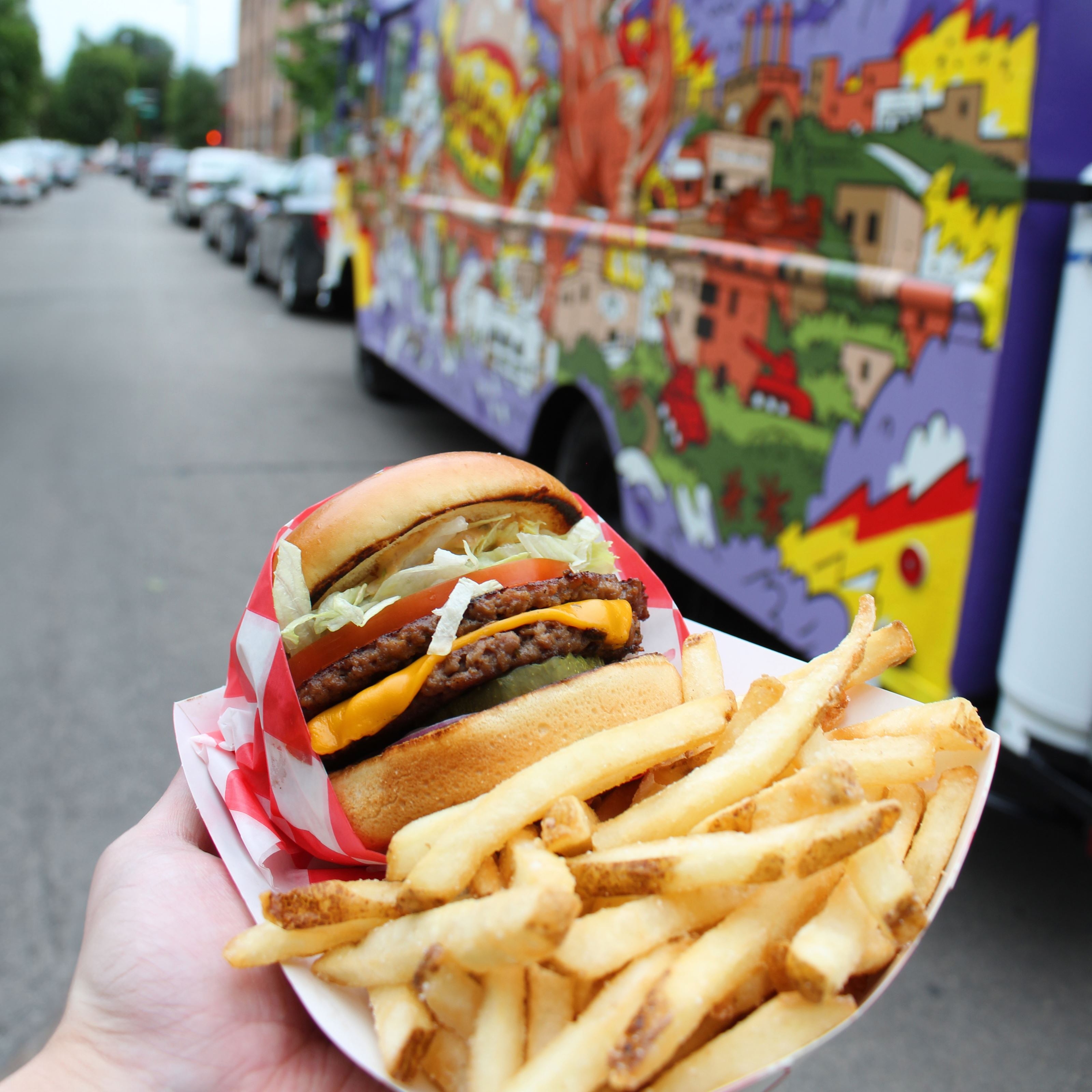 A hand holds out a paper tray containing a burger and french fries. In the background is a graphic, painted purple food truck.