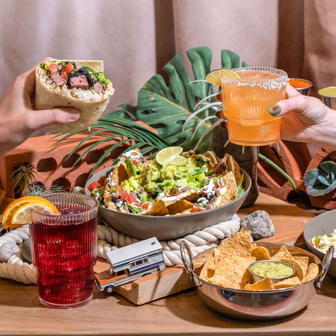 Two hands, one holding a burrito and the other holding a cocktail, reach into the frame where a table is displayed with another cocktail, a burrito bowl and a bowl of tortilla chips.