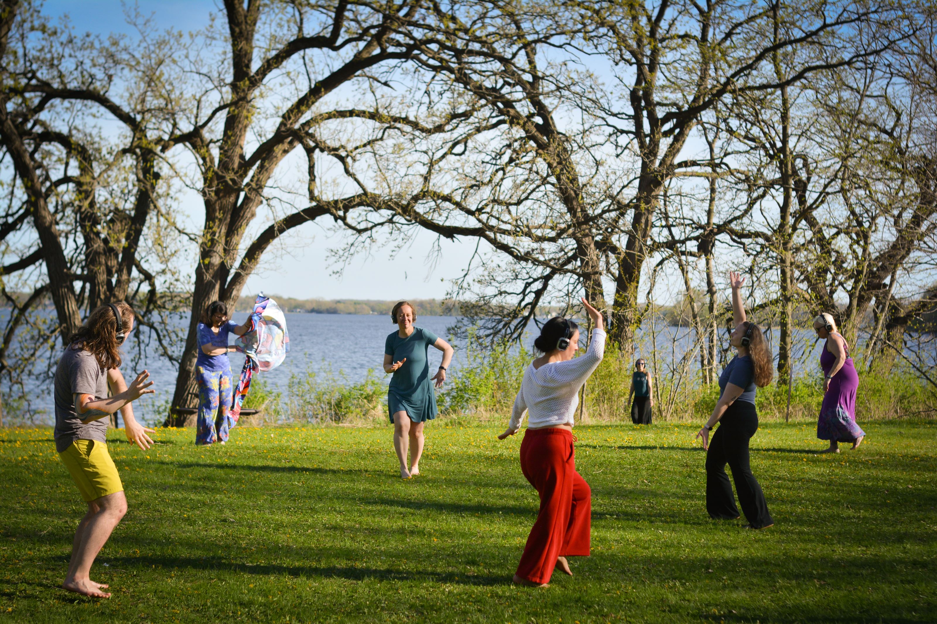 People appear in dance-like poses on a grassy area next to a lake.
