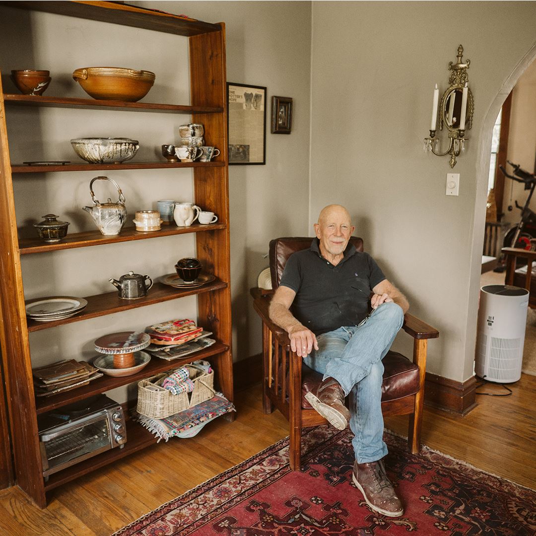 A man sits in a chair with pottery on shelves behind him.
